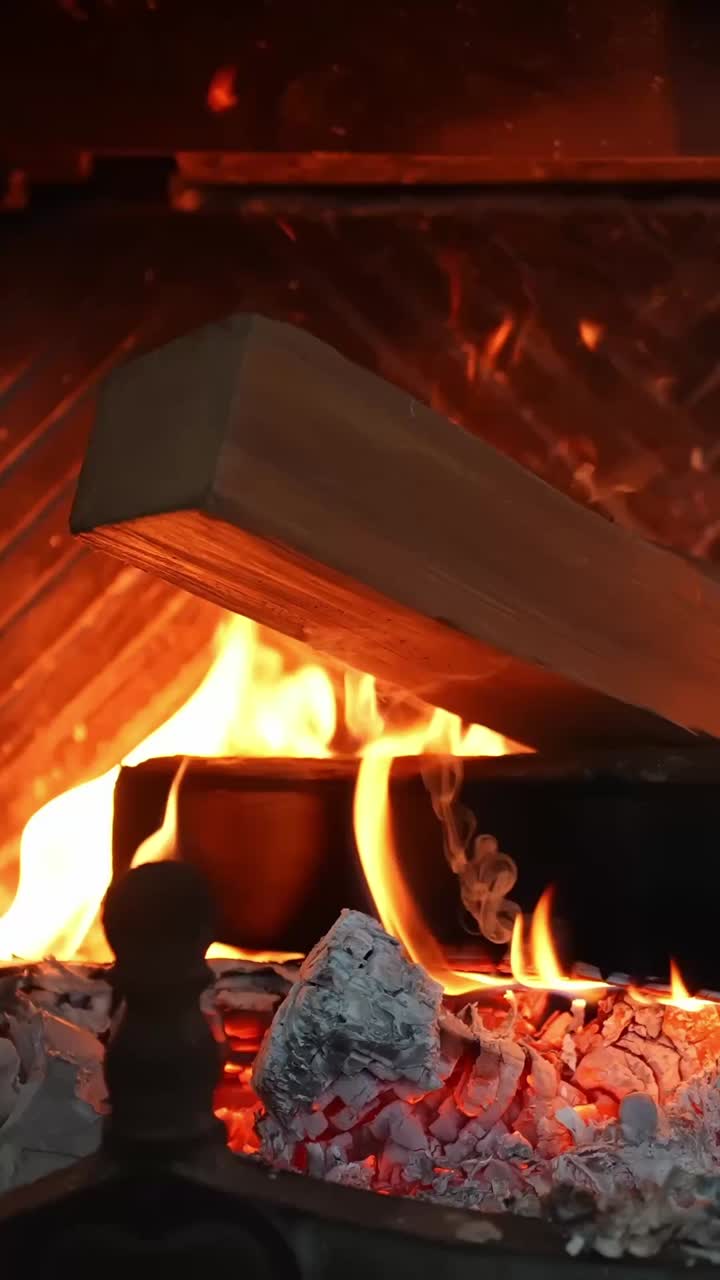 Vertical close-up inside a fireplace insert as a fresh log catches—bright flames, flying sparks, and glowing embers over ash and coals, creating cozy home heat