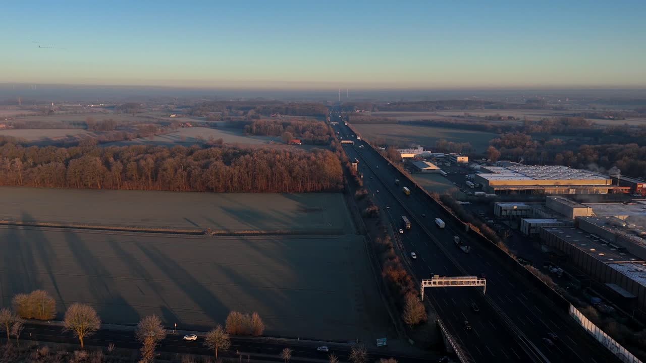 Busy traffic on german autobahn highway during frosty winter day at sunrise. Aerial drone wide shot. Farm fields and industrial warehouses and buildings in suburb. Forest landscape in USA.