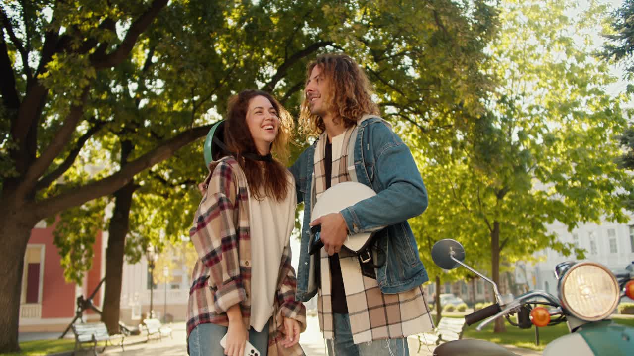 Happy couple, a guy with long curly hair in a denim shirt and his brunette girlfriend are dancing and hugging near their green moped in a summer city park