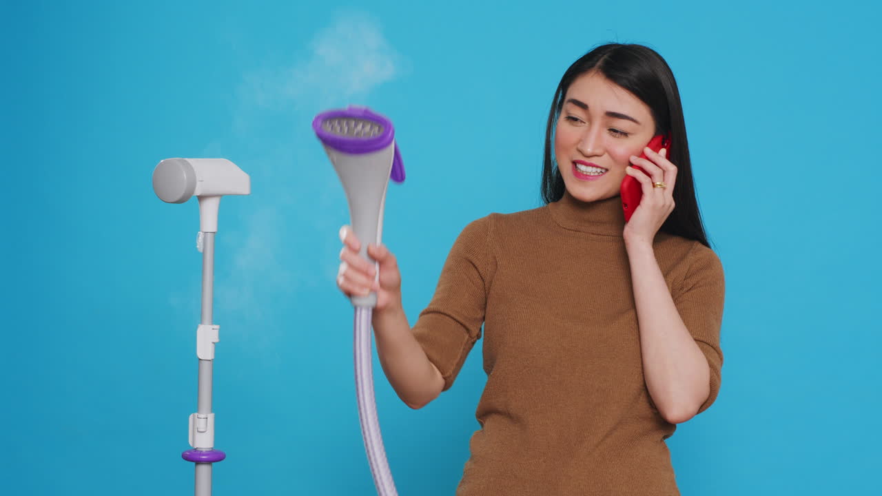 Cleaning lady talking at mobile phone with remote friend while using steamer to ironing clothes