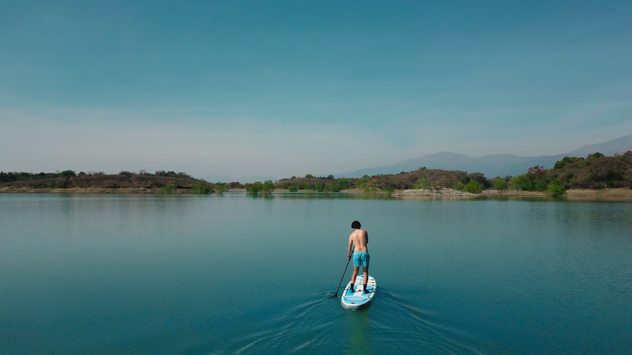 Pria Mendayung Papan Dayung di Danau Biru Tenang dengan Pegunungan di Latar Belakang