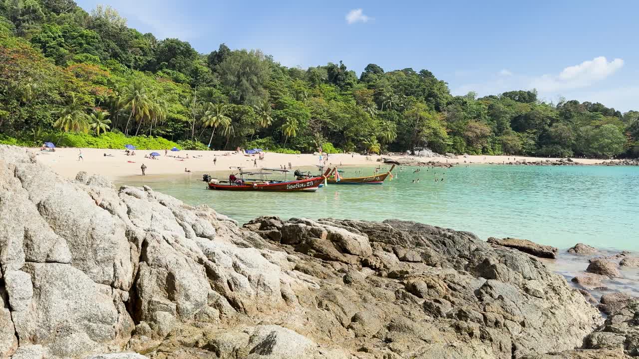 A tranquil beach in Phuket with turquoise waters, rocky foreground, and lush greenery under bright daylight