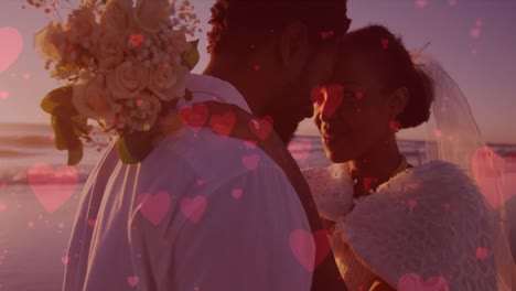 red hearts floating against african american newly married couple embracing each other at the beach