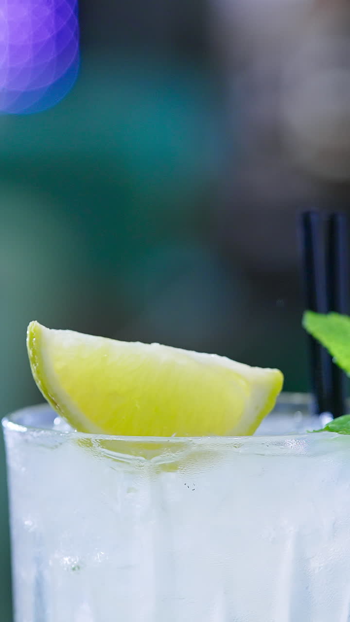 Upper part of glass filled with light beverage and two black straws in. Ice cubes, lemon slice and leaves of mint on top. Close up. Blurred backdrop. Vertical video