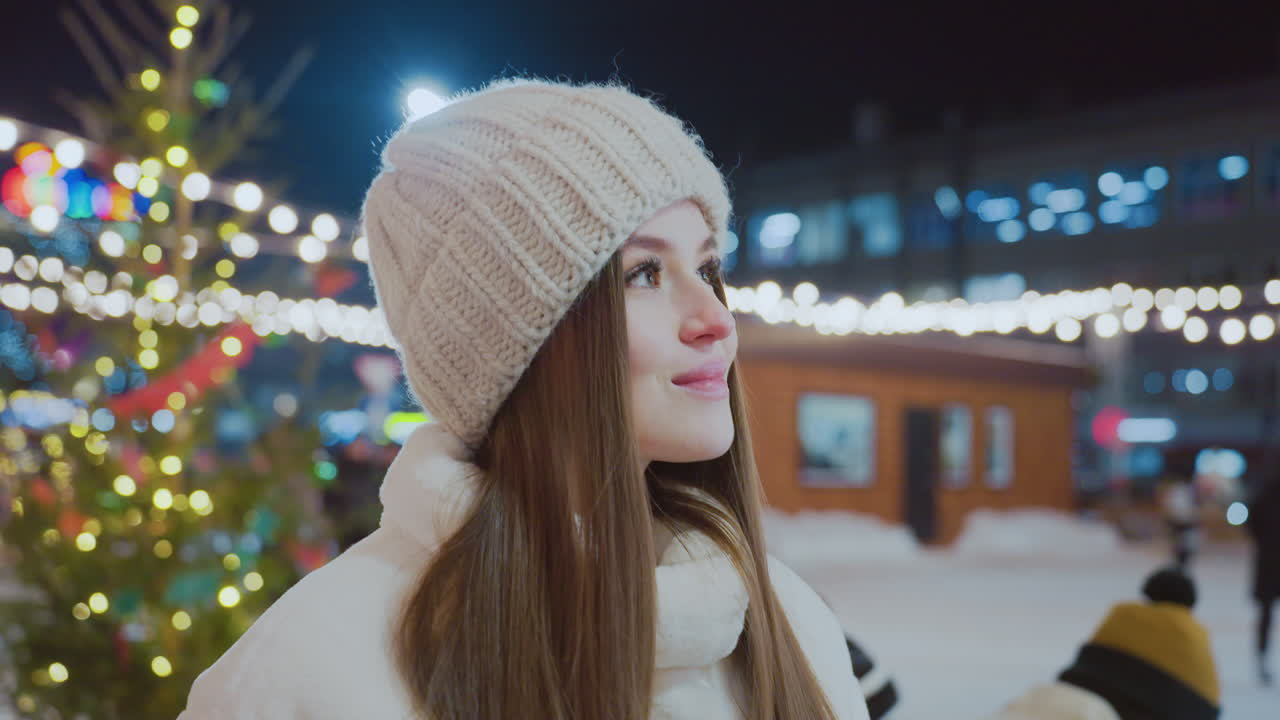 Smiling woman in warm winter outfit enjoys festive atmosphere at night, surrounded by twinkling lights, decorated Christmas tree, and people in the background