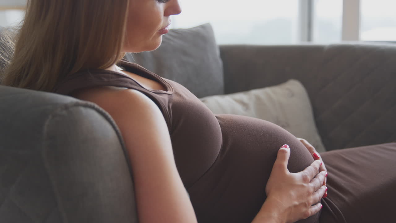 Pregnant Woman Relaxing On Sofa At Home Holding Stomach Feeling Baby Kick