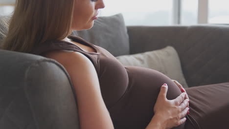 Mujer-Embarazada-Relajándose-En-El-Sofá-En-Casa-Sosteniendo-El-Estómago-Sintiendo-Una-Patada-De-Bebé