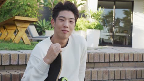 portrait of happy asian male teenager sitting with skateboard on sunny day in garden