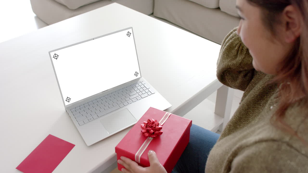 Caucasian woman holding gift and using laptop with copy space on blank screen