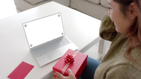 caucasian woman holding gift and using laptop with copy space on blank screen