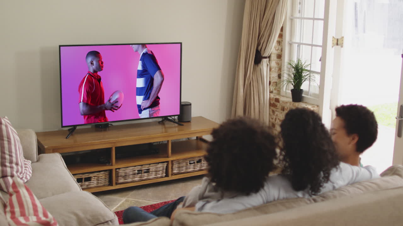 familia biracial viendo la televisión con diversos jugadores de rugby masculinos con la pelota en la pantalla
