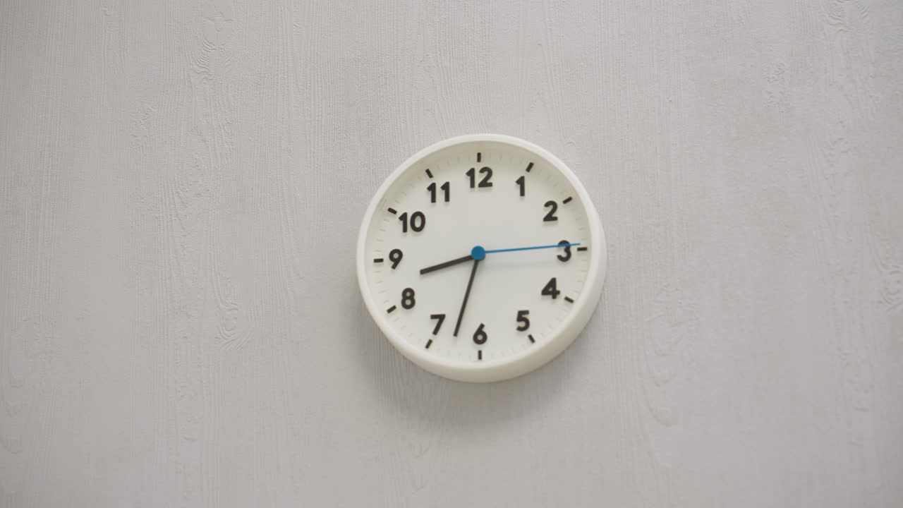 White round wall clock with black numbers and hands, blue second hand moving continuously, mounted on light gray textured wall in minimalist indoor space, evoking calm and passage of time
