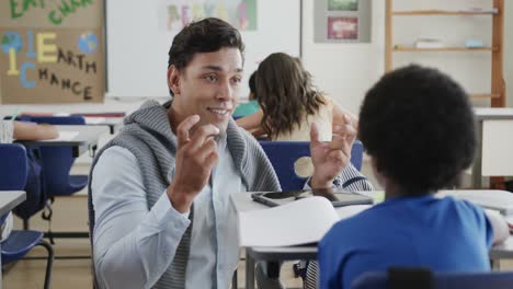 happy diverse male teacher talking to boy at desk in elementary school class, slow motion