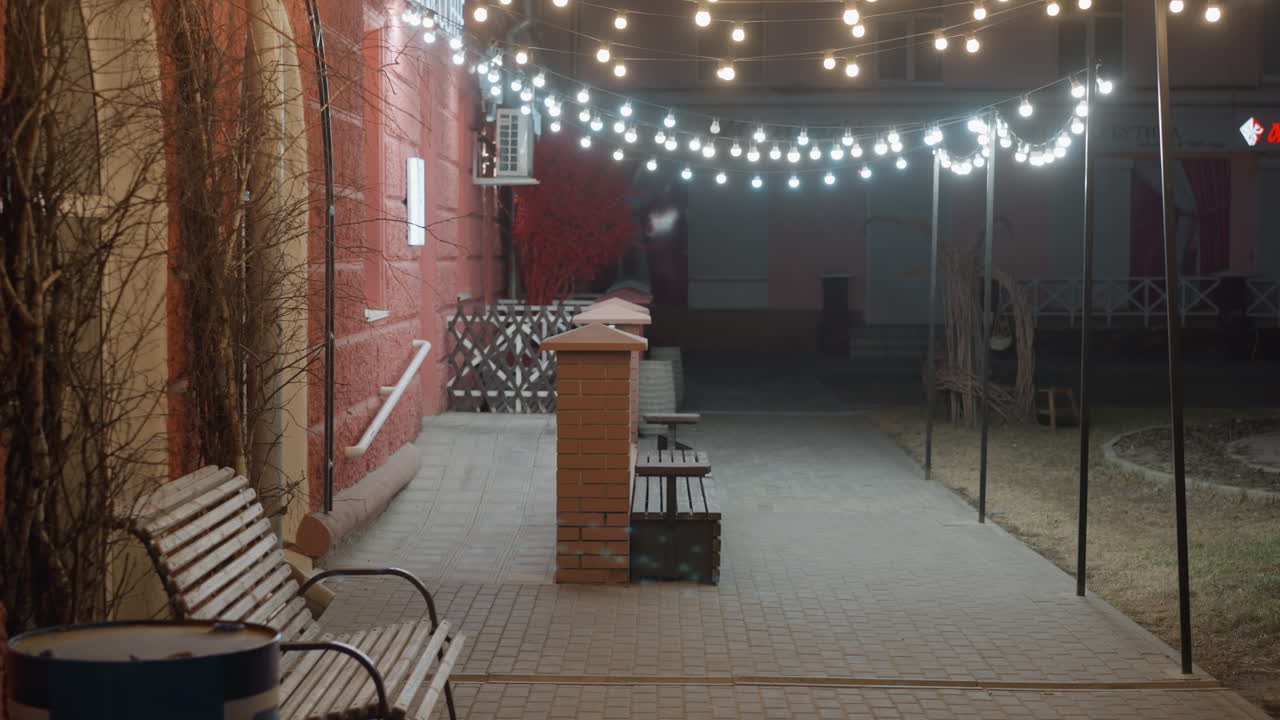 A cozy evening outdoor scene with string lights illuminating a pathway lined with benches and a brick wall