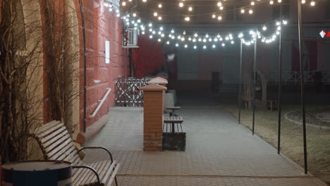 a cozy evening outdoor scene with string lights illuminating a pathway lined with benches and a brick wall