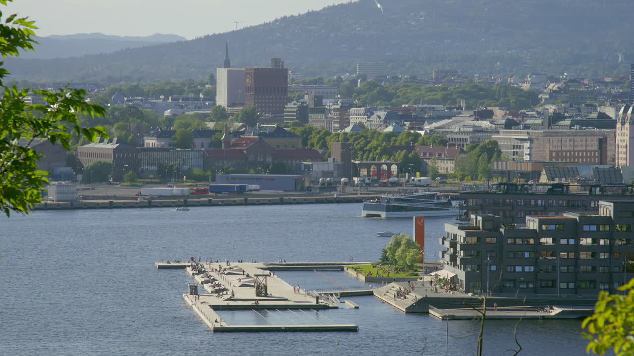 Scenic view of Oslo harbor with waterfront buildings, people enjoying the swimming aread, and a boat on calm water.