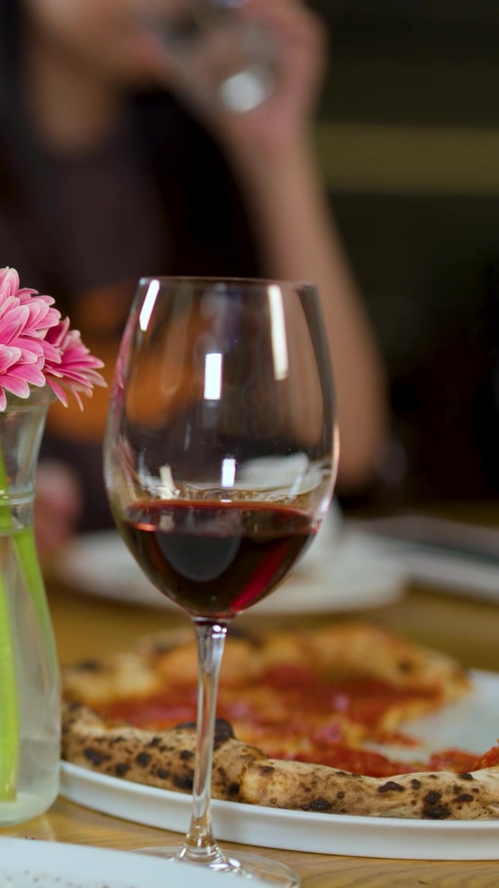 Primo piano di un bicchiere di vino rosso e pizza sul tavolo con amici che cenano sullo sfondo.