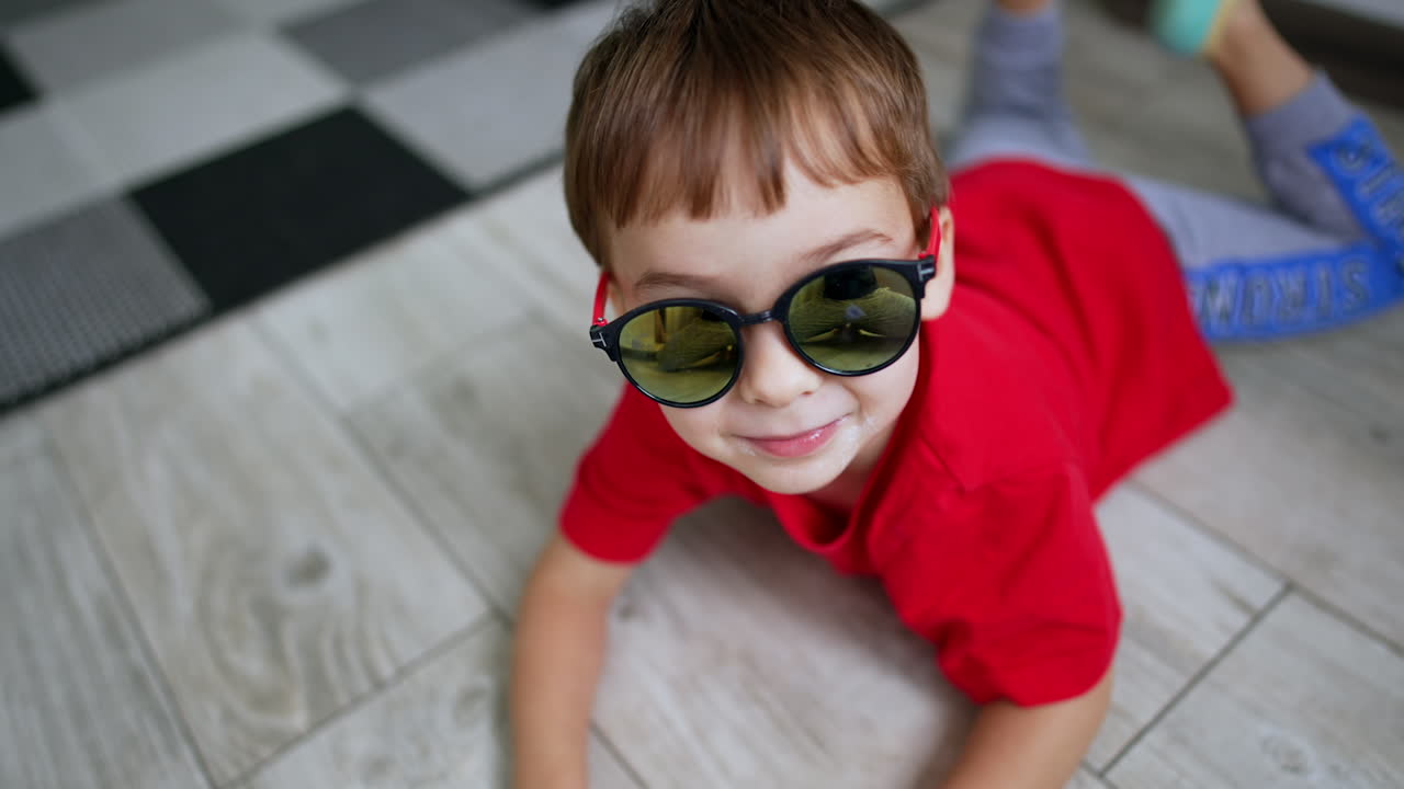 Cute Caucasian toddler wearing sunglasses lies on the floor. Happy kid having fun playing at home. Close up.