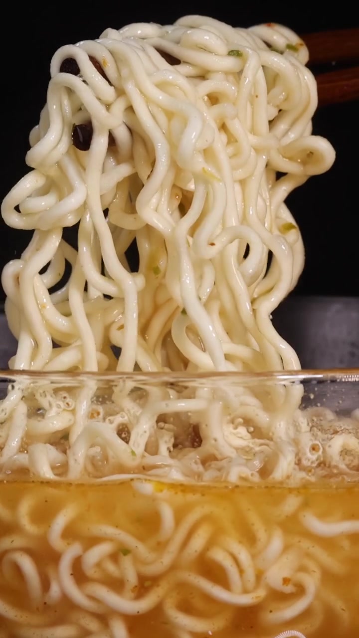 黒い背景をバックに、ガラスの器から湯気の立つインスタントラーメンを木製の箸で持ち上げるクローズアップ。