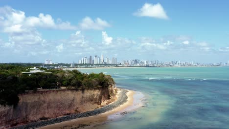 Trucking-right-aerial-drone-shot-of-the-tropical-beach-capital-city-of-Joao-Pessoa,-Paraiba,-Brazil-from-the-touristy-Cabo-Branco-cliffs-with-stunning-clear-turquoise-ocean-water-on-a-summer-day