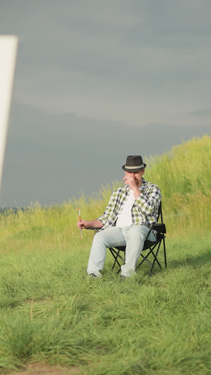 In a sunlit meadow, a focused female artist in a white dress paints on a canvas with a palette in hand, while a man in a black hat and checkered shirt sits calmly in a chair, observing the scene