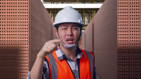 close up of angry asian male engineer with safety helmet shouting at someone while standing with red brick packed in stacks are stored