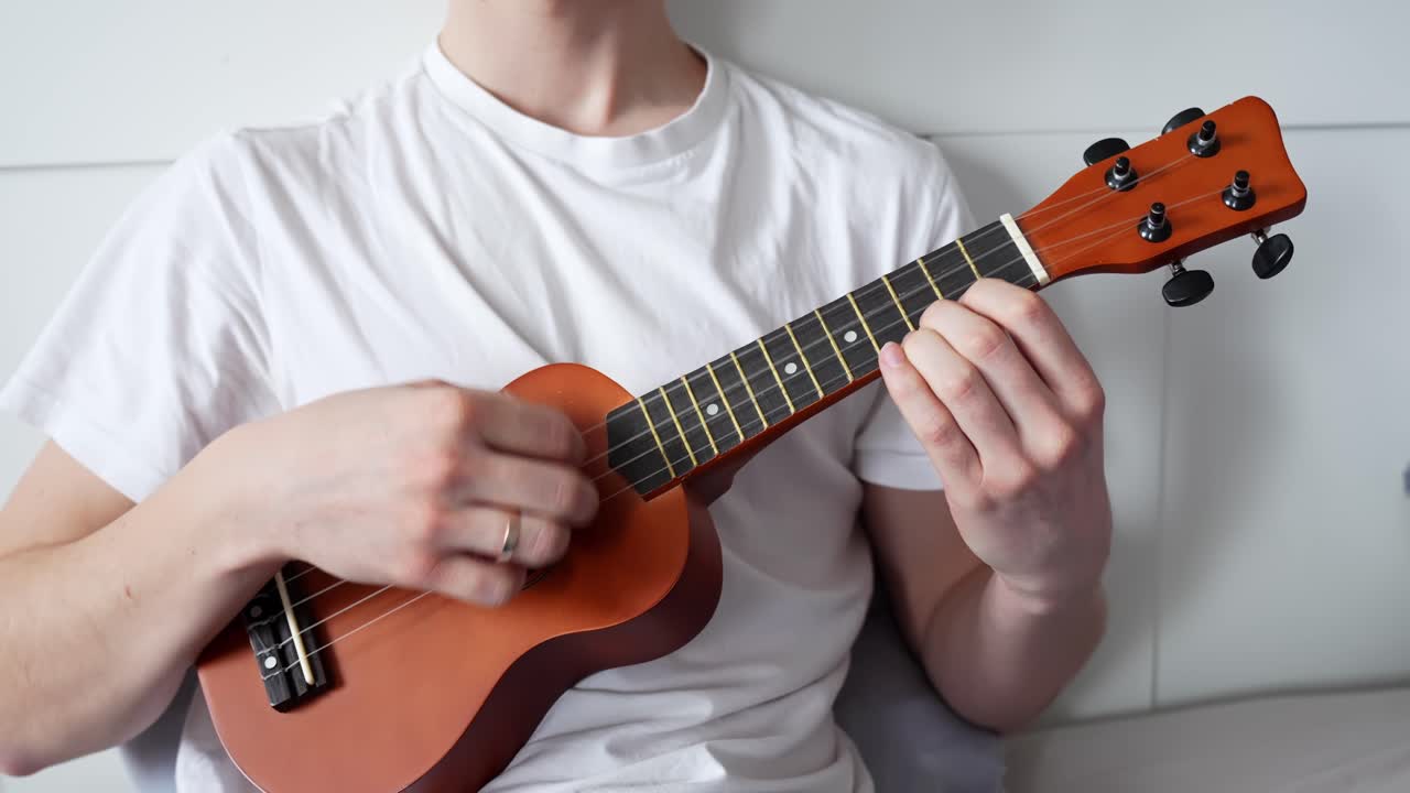 A man dressed in a plain white t-shirt plays a wooden ukulele while seated indoors, capturing his hand positions on the strings, showcasing casual music-making in a relaxed setting.