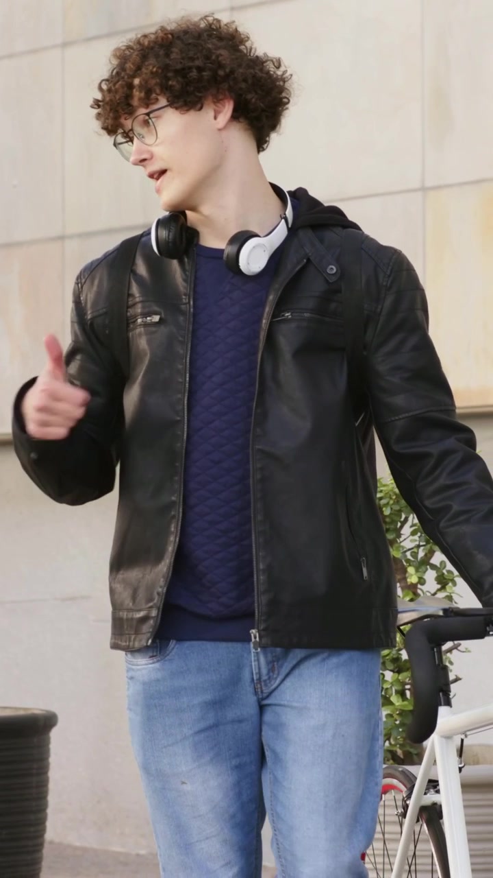 Vertical video: Stepping leather-jacketed man guiding bike on sidewalk gesturing at passerby
