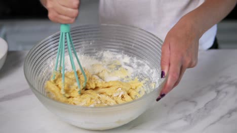 Vista-De-Cerca-De-Las-Manos-Femeninas-Mezclando-Masa-Pegajosa-En-La-Cocina-Usando-Un-Batidor.-Comida-Hecha-En-Casa.-Tiro-En-Cámara-Lenta