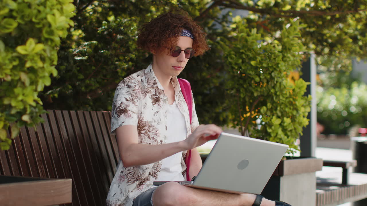 Happy young man working online distant job with laptop in city sunshine street sitting on bench