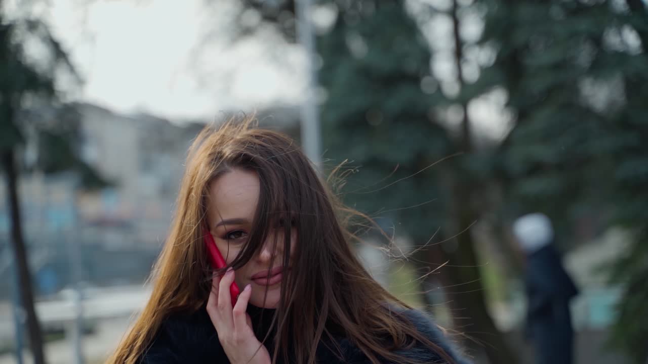 Portrait of a beautiful woman with long hair. Dark-haired lady talking on the phone and posing to camera on a city background. Slow motion.