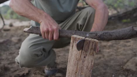 Caucasian-male-survivalist-using-branch-and-machete-to-cleave-firewood-at-camp-in-wilderness