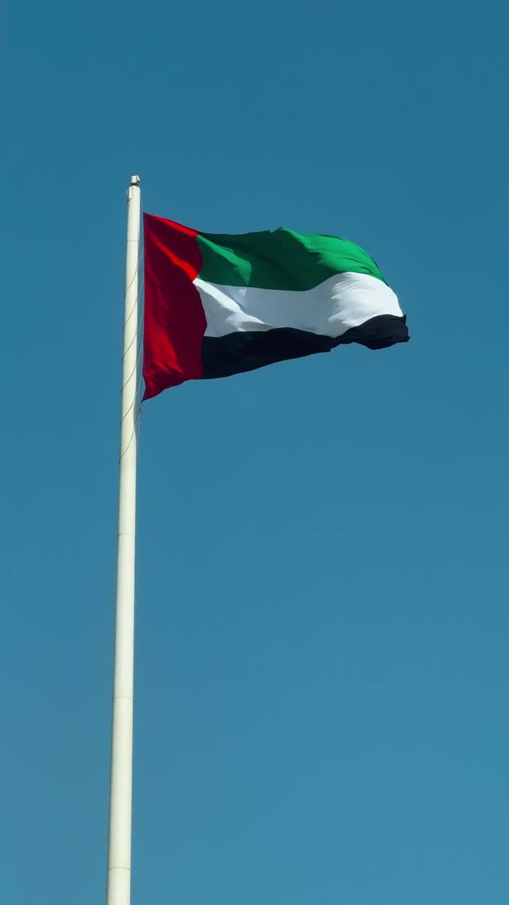 A close-up shot of the UAE national flag waving boldly atop a tall flagpole, set against a clear blue sky near Abu Dhabi, capturing national pride, color, and movement in a bright, serene atmosphere
