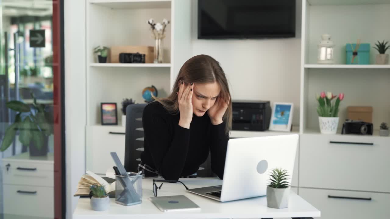 The young woman in glasses is sitting at a desk in an office in front of a laptop