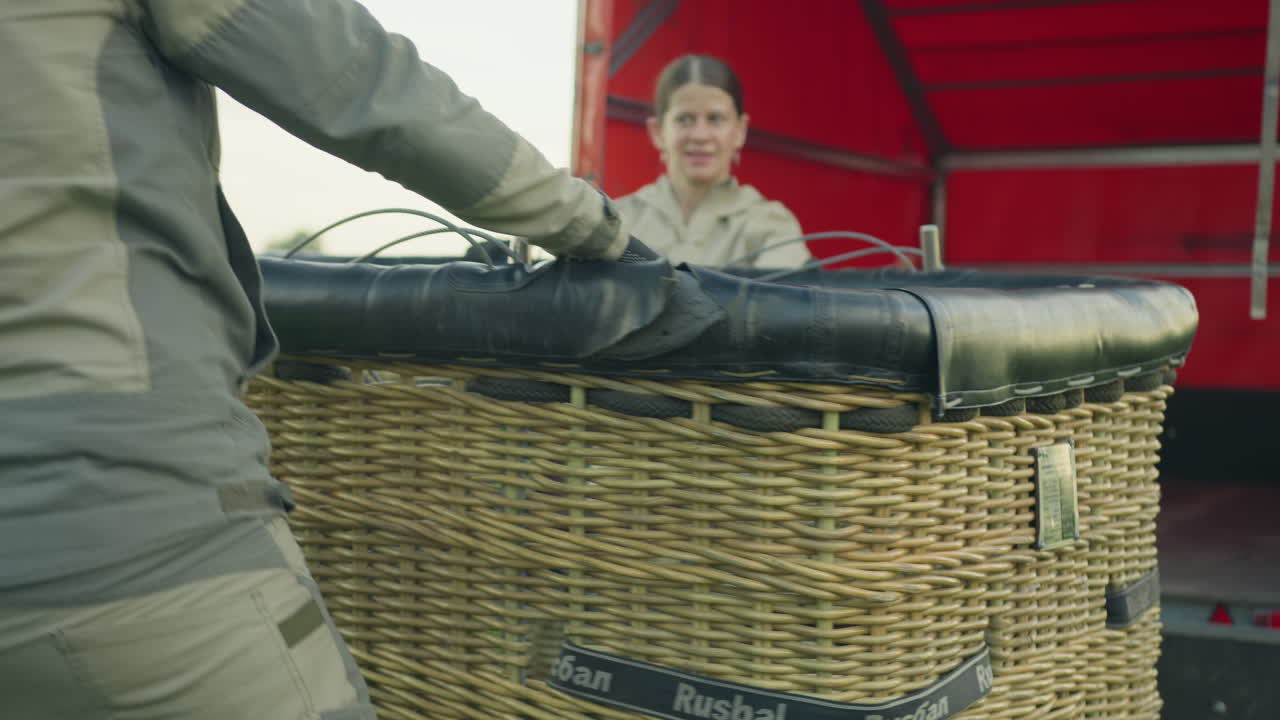 Man and woman in workwear unload massive hot air balloon basket from back of truck with red canopy, coordinating effort with focus and teamwork in grassy rural setting