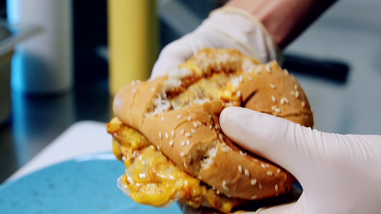 Lækker fastfood. En kok deler en frisk burger i to. En stor, appetitlig cheeseburger med bøf, smeltet ost, grøntsager og sauce i kokkens hænder.