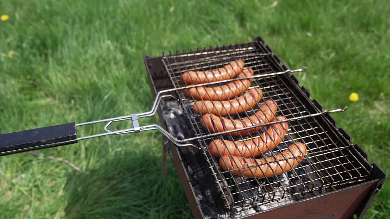 Tasty juicy sausages grilling over a fire in smoke. Hot dog sausages on a grill grate cooking on the green grass background. Barbeque in summer.
