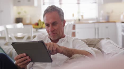happy caucasian man sitting on sofa in living room, using tablet