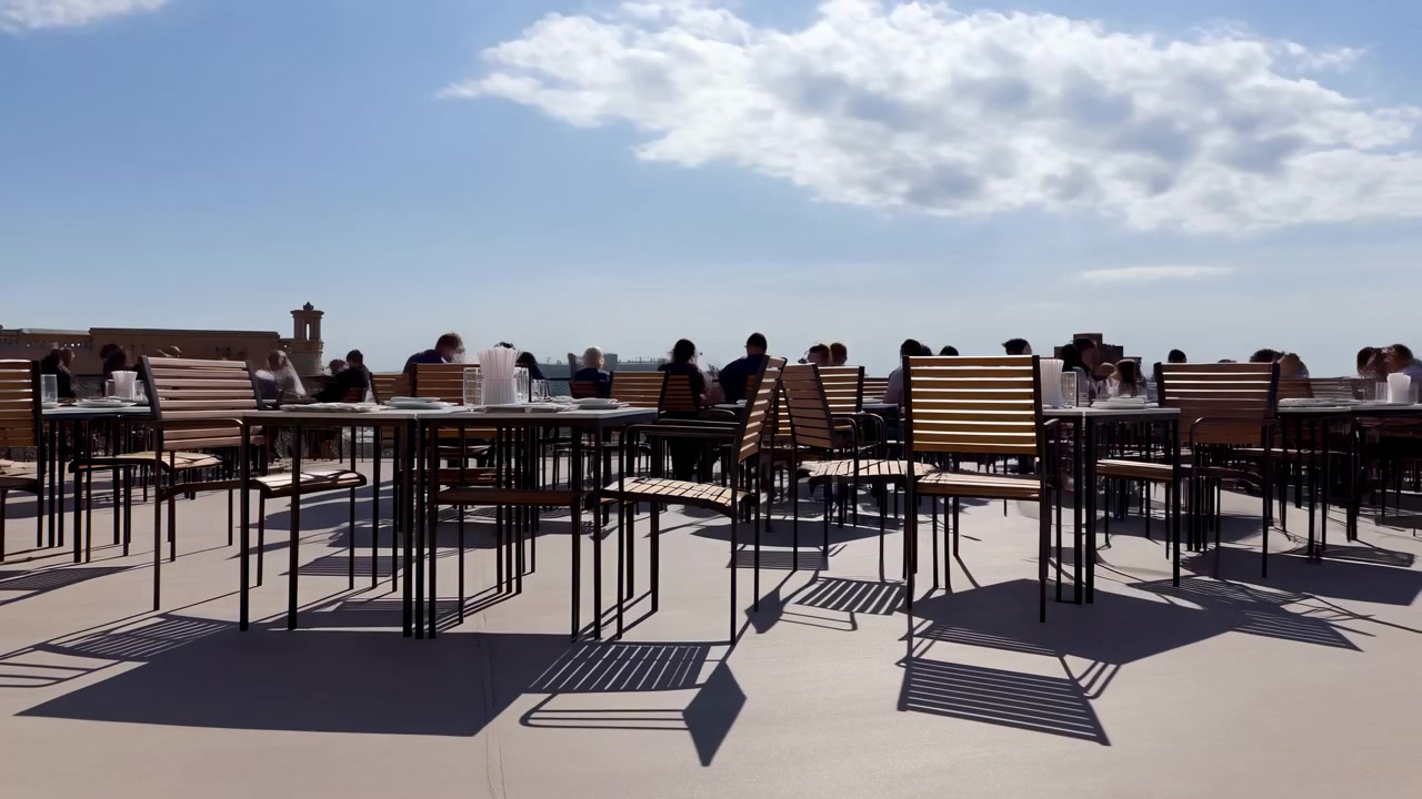 File di tavoli da esterno e sedie a doghe si riempiono mentre le persone arrivano e si siedono su una luminosa terrazza all'aperto, con bicchieri, viste sulla città, nuvole alla deriva e ombre direzionali nette