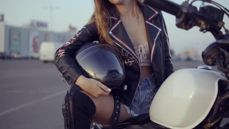 pretty female biker in shorts sitting on the motorcycle seat and holding her helmet. young curly woman in sunglasses looking in