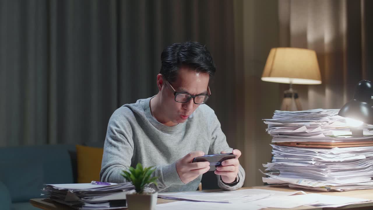 Un uomo asiatico si diverte a giocare a un gioco sullo smartphone dopo aver lavorato con documenti a casa.