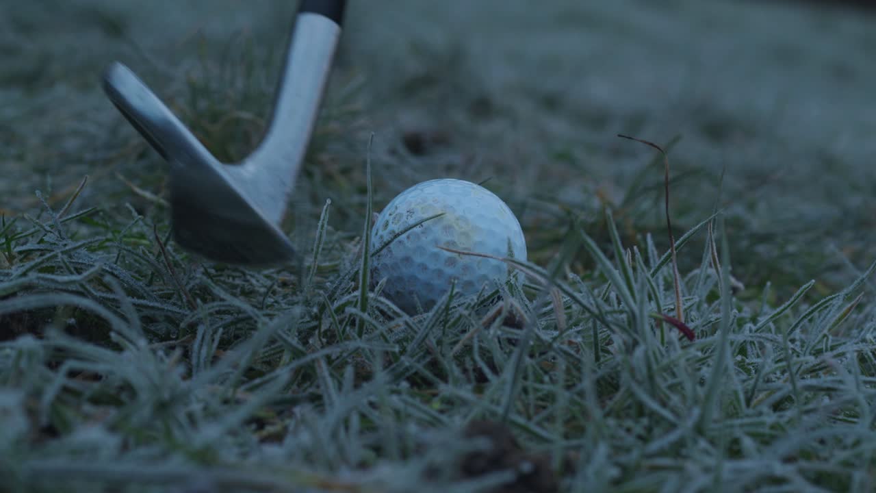 Close-up static shot of a golf ball in frosted grass being hit by golf club during early morning game