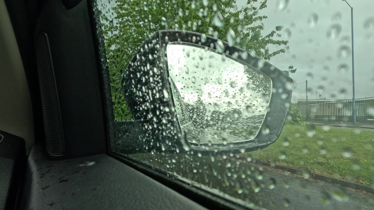 Vista suggestiva dall'interno di un'auto parcheggiata durante una giornata di pioggia in Inghilterra. Le gocce di pioggia coprono il finestrino laterale e lo specchietto retrovisore, mentre il cielo coperto e le strade bagnate creano una tipica scena del tempo britannico.