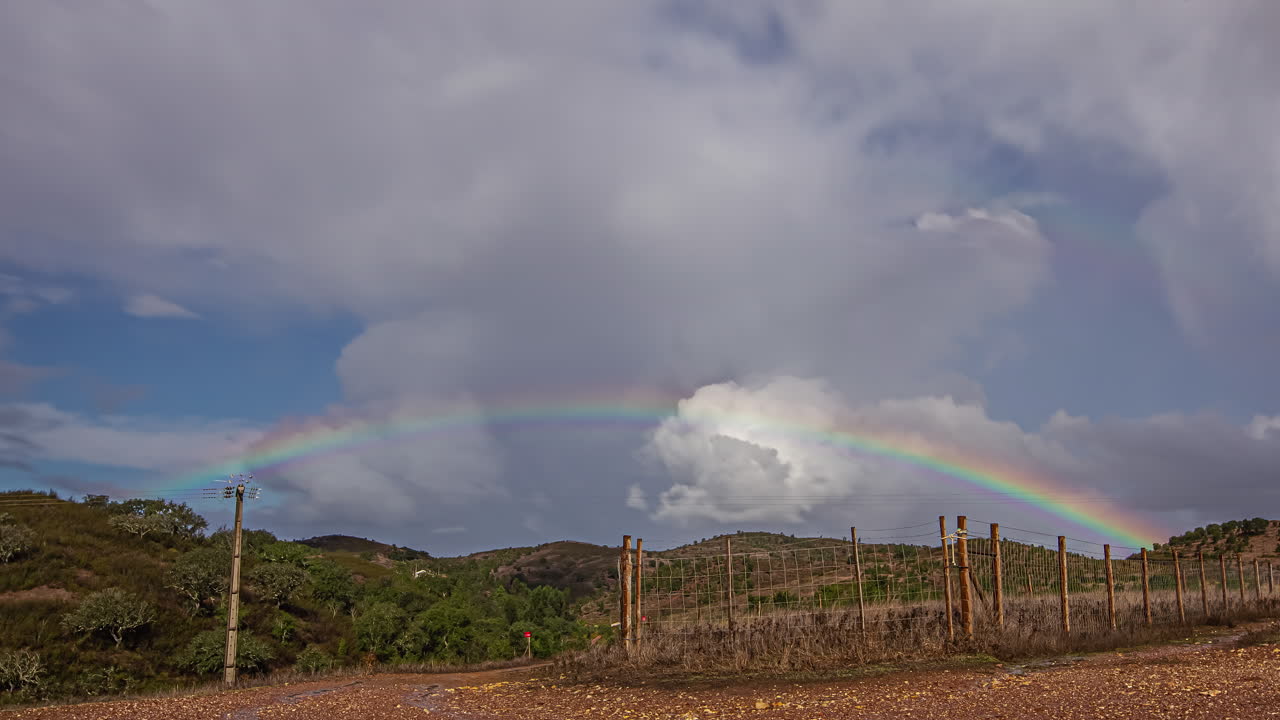 cielo azul con arco iris y nubes blancas moviéndose sobre un campo seco con cercas