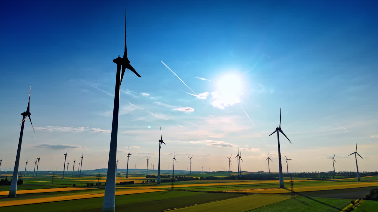 Wind turbines in a sunny field landscape. Several wind turbines spin in a vast green field under a bright blue sky with scattered clouds