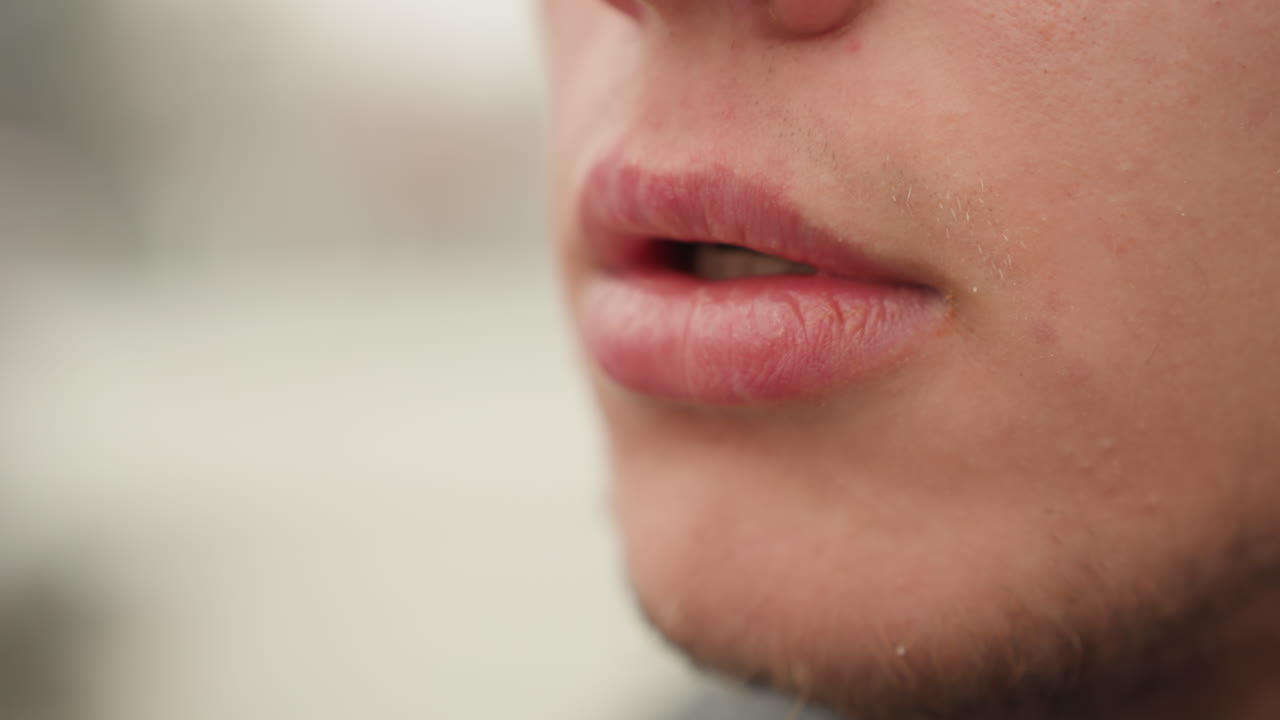 Primo piano delle labbra di un uomo che esalano fumo di sigaretta, mano guantata vicino alla bocca, cenere incandescente sulla punta, sfondo sfocato, respiro freddo visibile, dettaglio della barba corta, inquadratura cinematografica sensuale e gradazione di colore tenue.