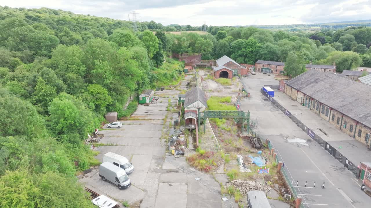 Aerial view of a disused railway and industrial site at Elsecar Heritage, showing long pitched‑roof buildings, cracked paved yard with weeds, fenced sections and dense surrounding woodland