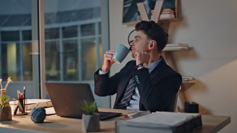 relaxed manager drinking coffee at laptop work desk. serious worker at evening