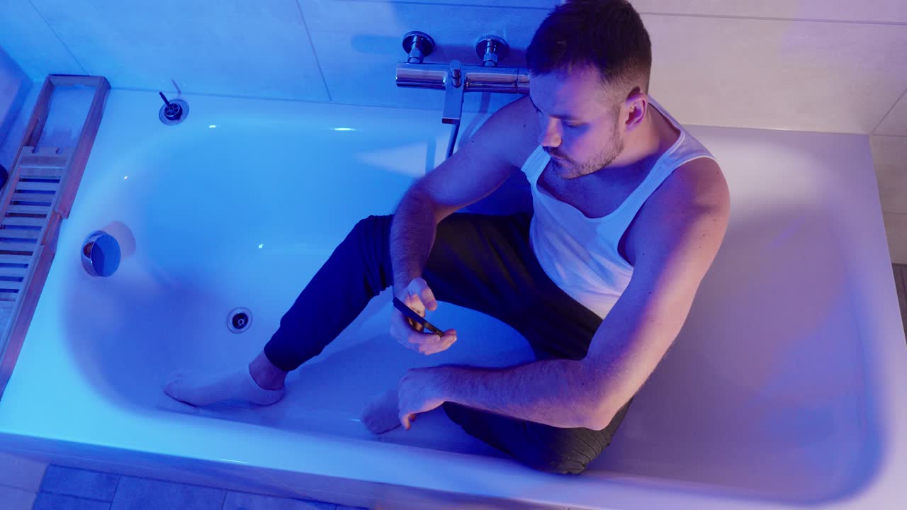 Cinematic shot of an emotional man sitting in a bathtub under blue ambient light, looking at his smartphone and shaking his head in sadness, symbolizing bad news, disappointment, digital stress