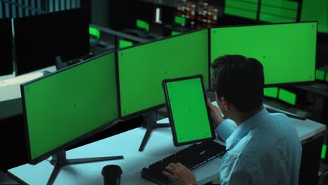vista lateral de un hombre asiático trabajando con una tableta y varios monitores de computadora con una pantalla verde simulada en la oficina